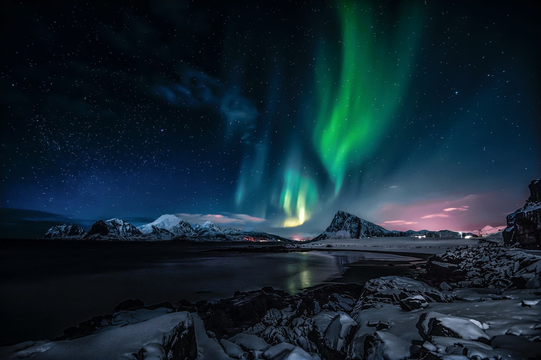 Green aurora over snowy mountains and a rocky shore on a starry night