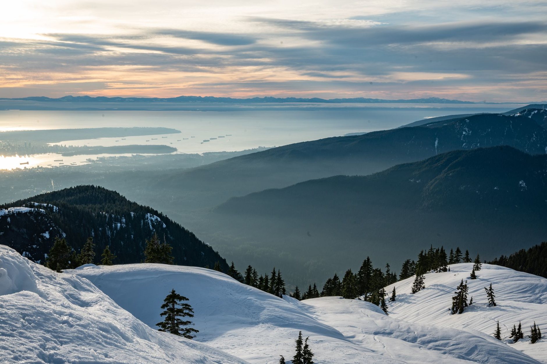 Snowy slopes dotted with small firs above misty valleys and the ocean at dusk
