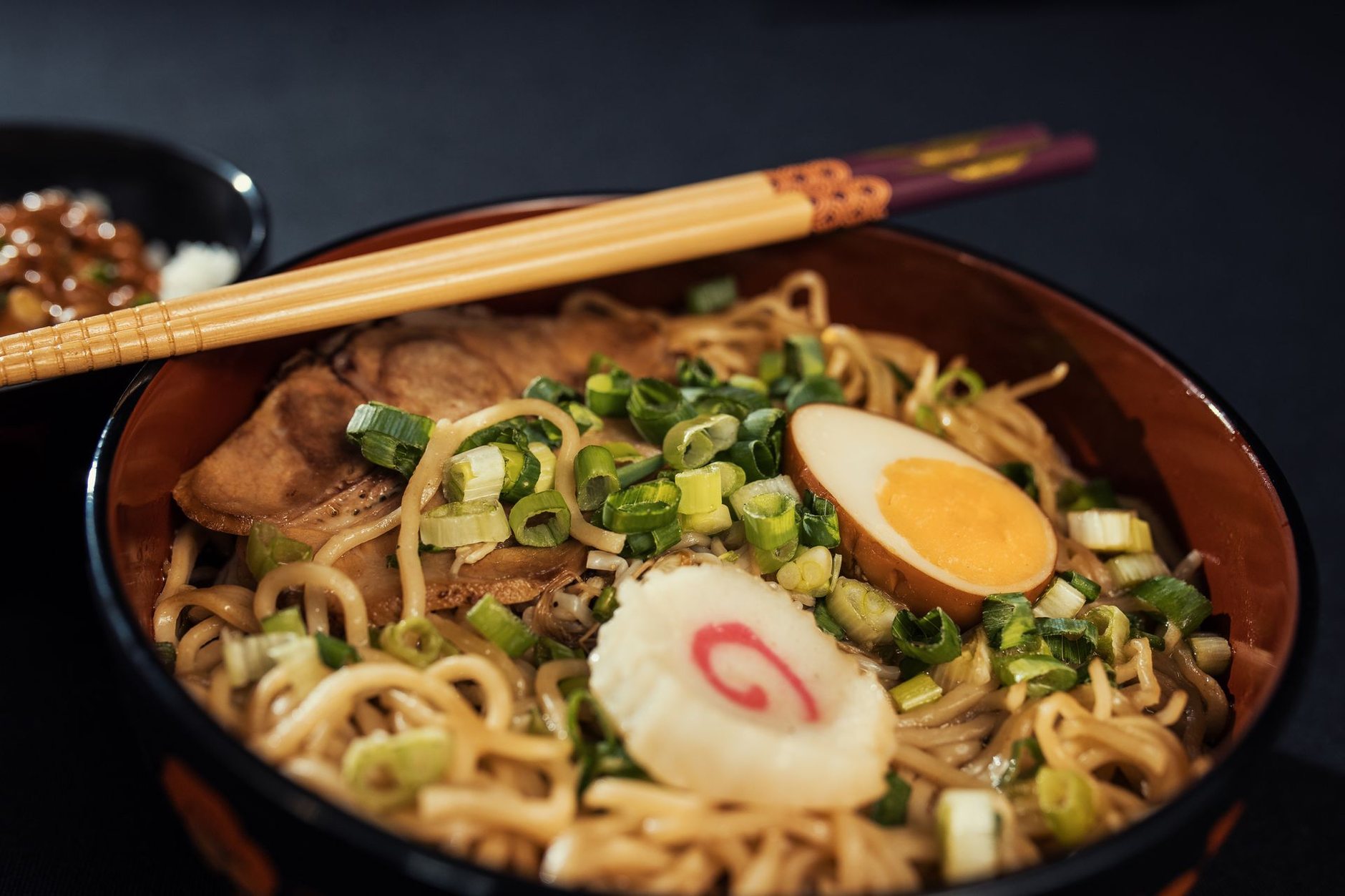 A bowl of ramen with chashu, a soft-boiled egg, narutomaki and green onion, chopsticks resting on the rim