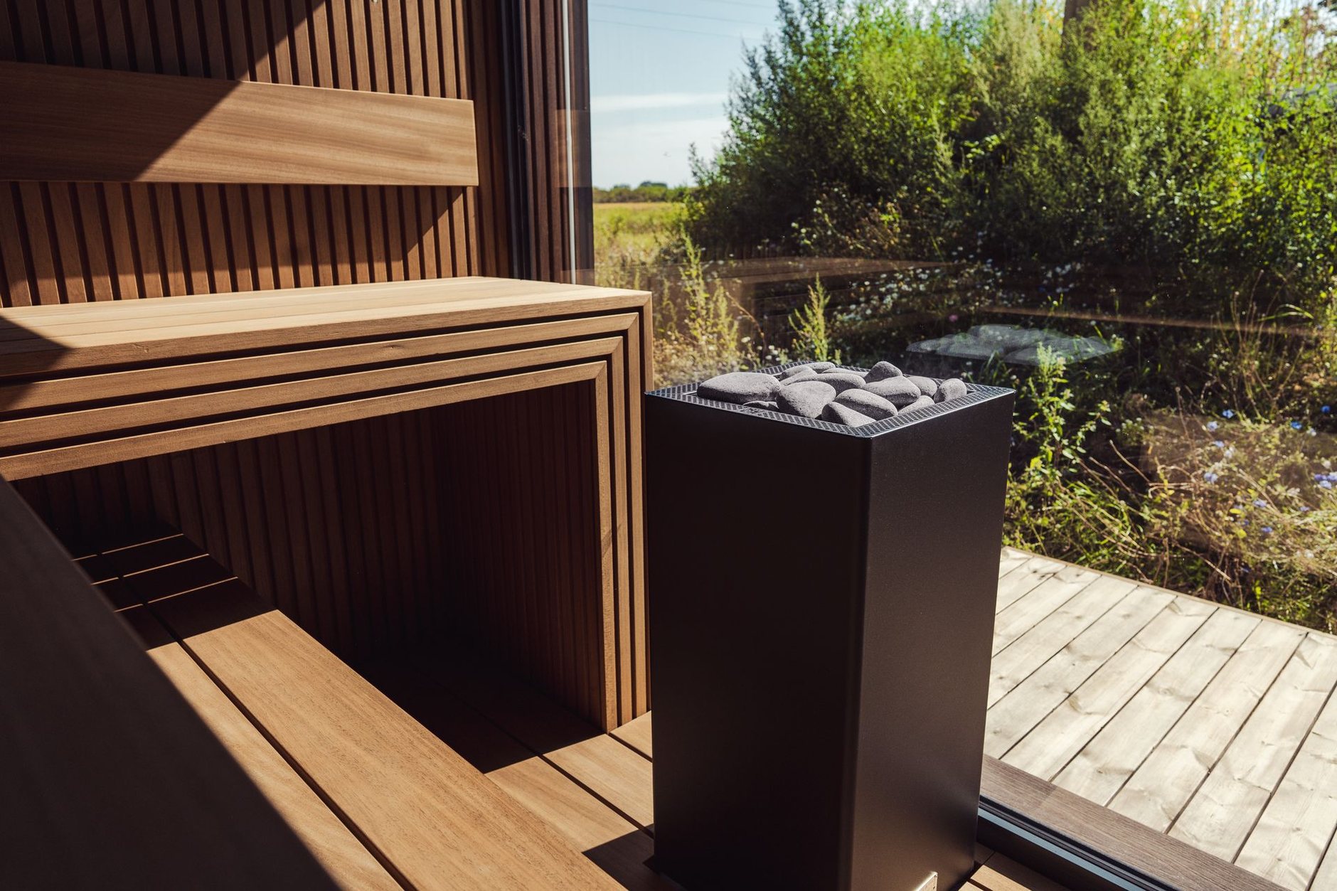 A modern wooden sauna with a black stone-topped stove and a glass wall facing greenery