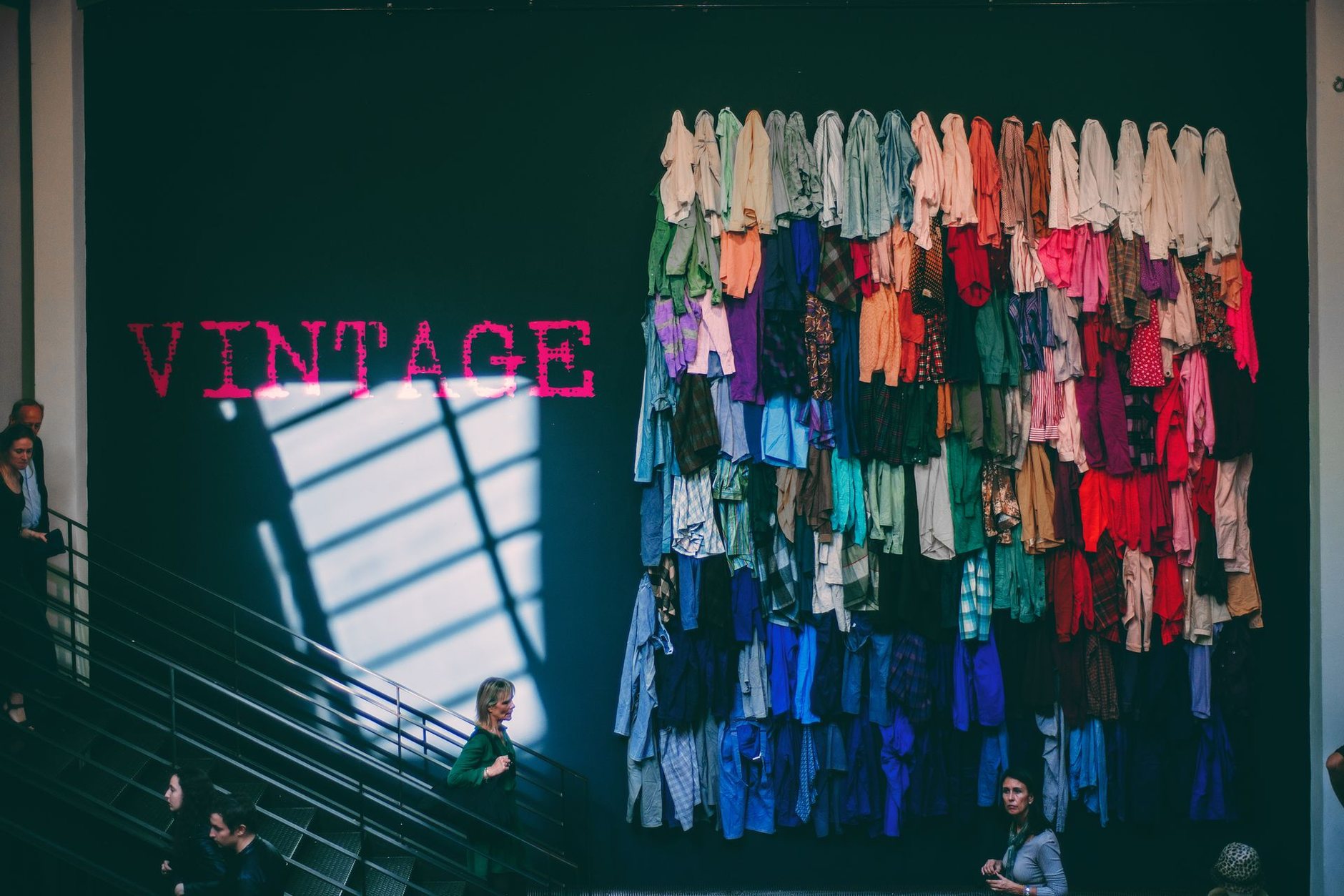 A wall hung with colourful clothes beside a red VINTAGE sign, with people on a staircase