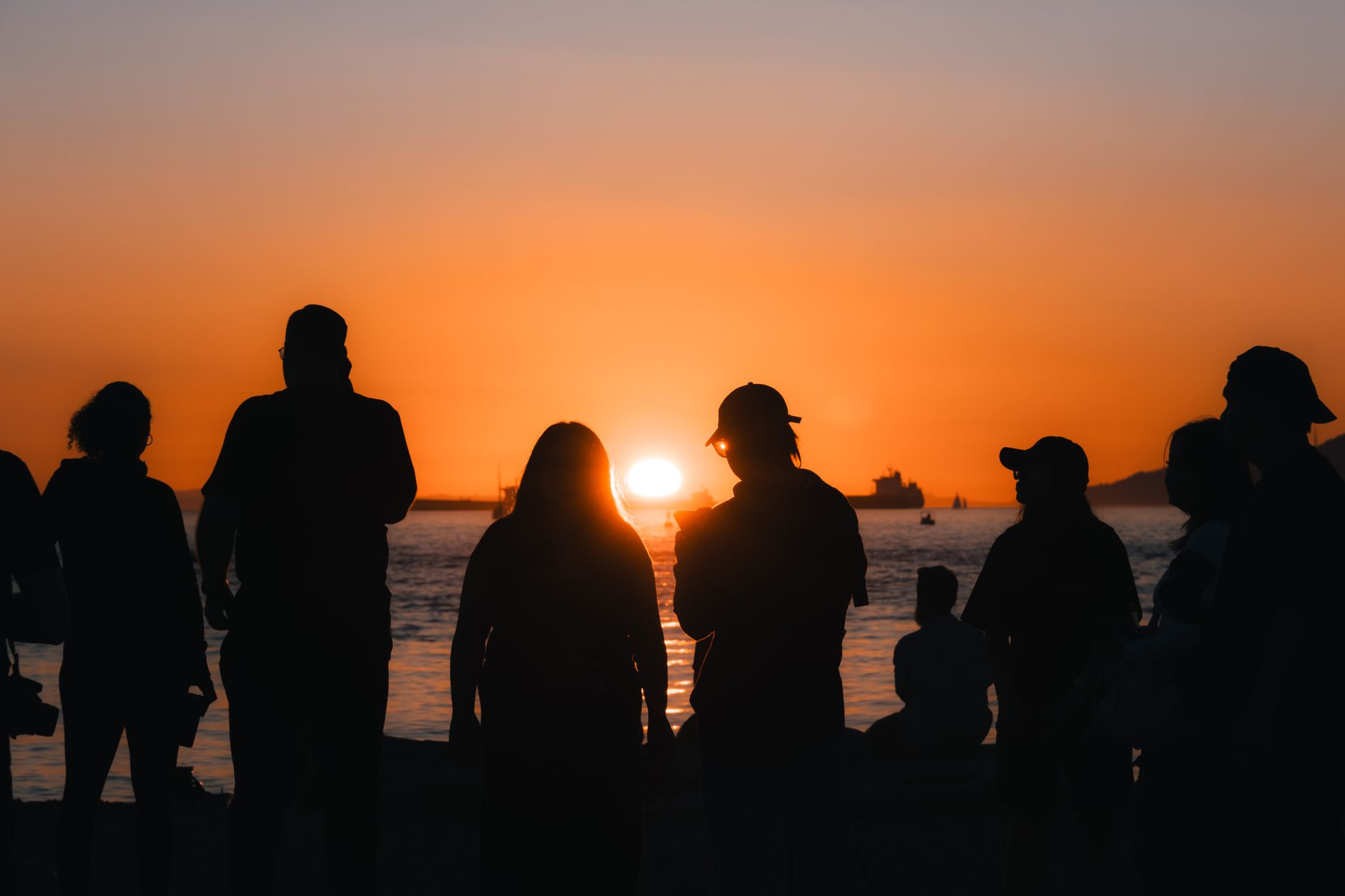 People silhouetted against a sunset over the water, with ships anchored offshore