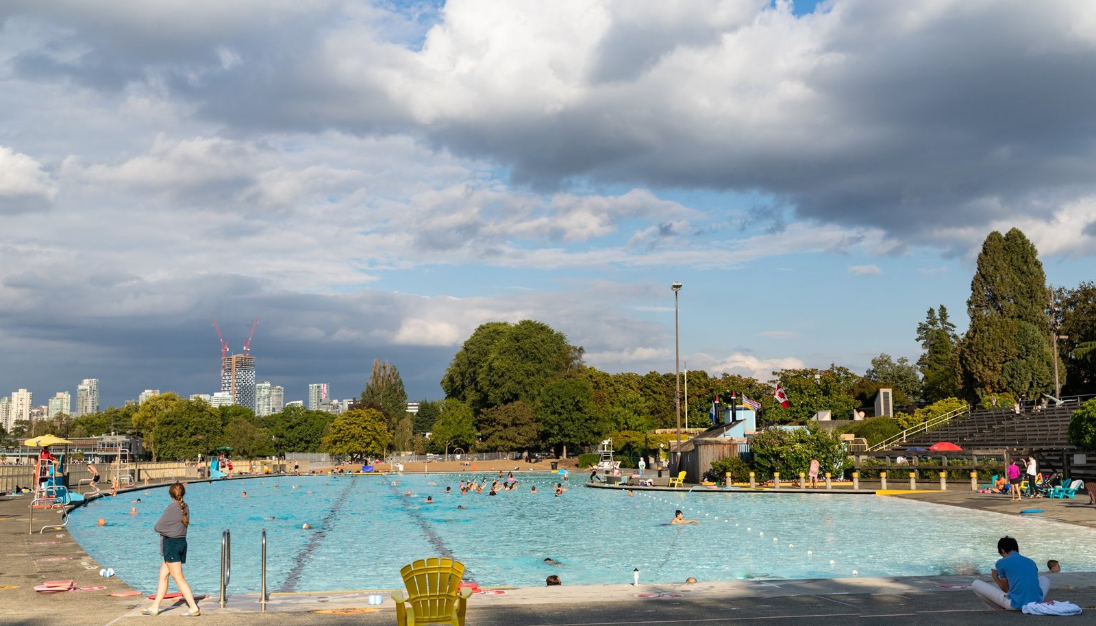 Swimmers in a long outdoor pool beside a leafy park, with downtown towers in the distance