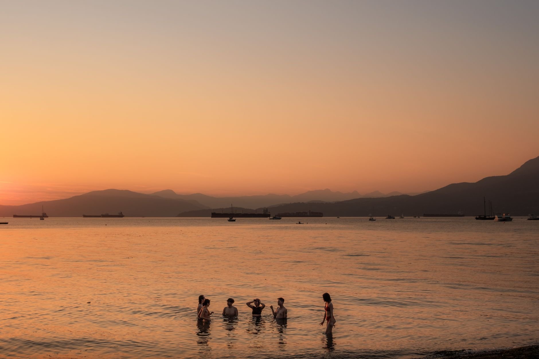 Bañistas entrando al mar al atardecer, con cargueros fondeados a lo lejos