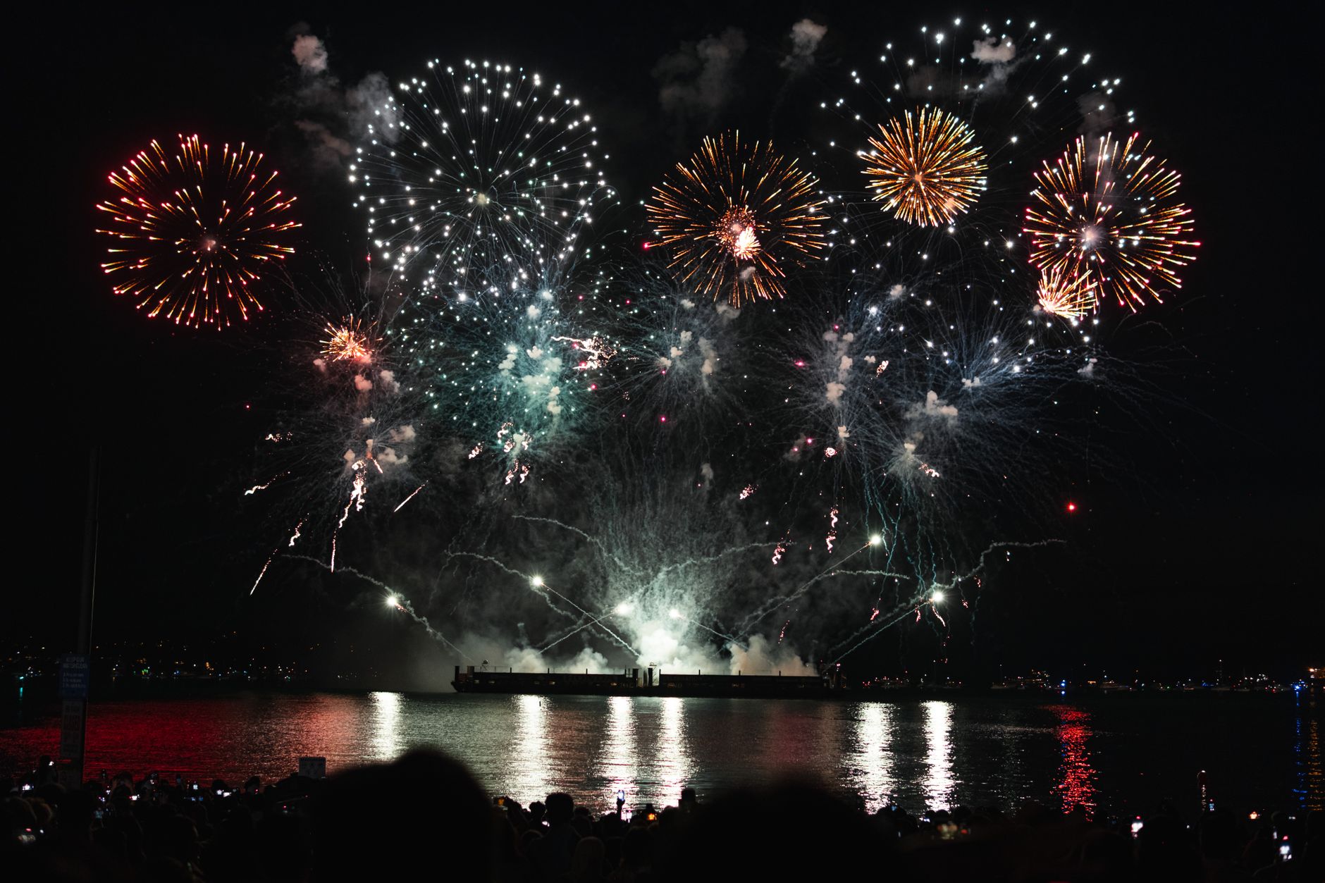 Fireworks bursting above a barge on the water at night, reflected in the bay